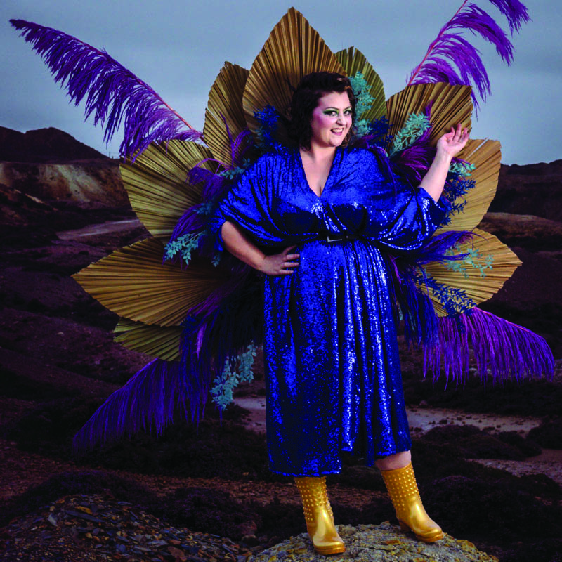 Kiri Pritchard-McLean in a sparkly blue dress and gold wellies with peacock feathers behind her.