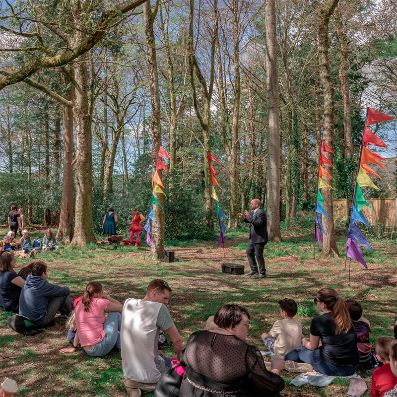 Photo of a man performing to a crowd outside in amongst some trees