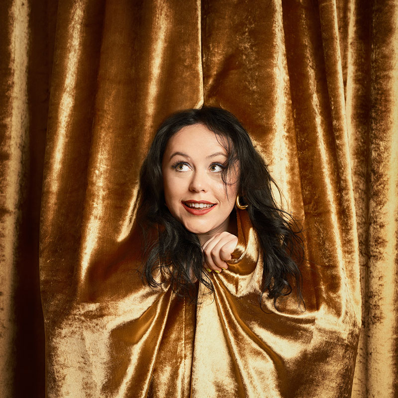 Photo of Amy Anette peaking out from between full length gold curtains