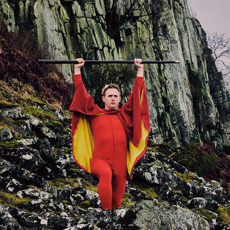 Photo of Chris Cantrill outside by some rocks dressed in red with a red cape holding a staff above his head