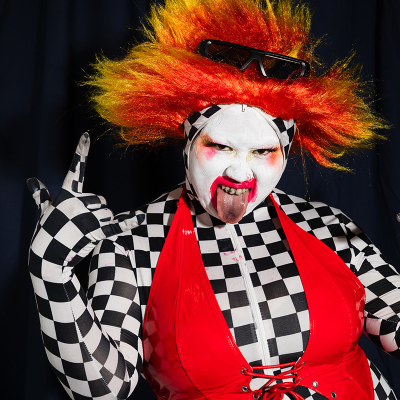 Su Mi in a fiery red and ornage wig, clown like make up and a checked body suit with a red dress over the top