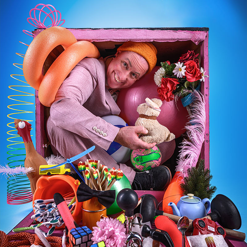 Photo of Daryll Carrington crammed into a box with lots of toys and props spilling out onto the floor