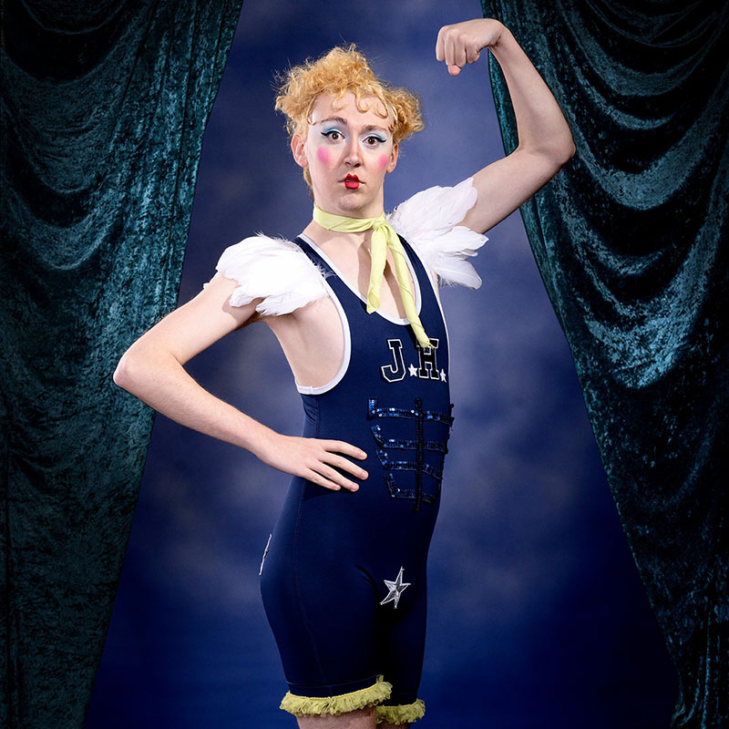 Photo of Lachlan Werner in a gym leotard with white feathers under the shoulder straps flexing a bicep