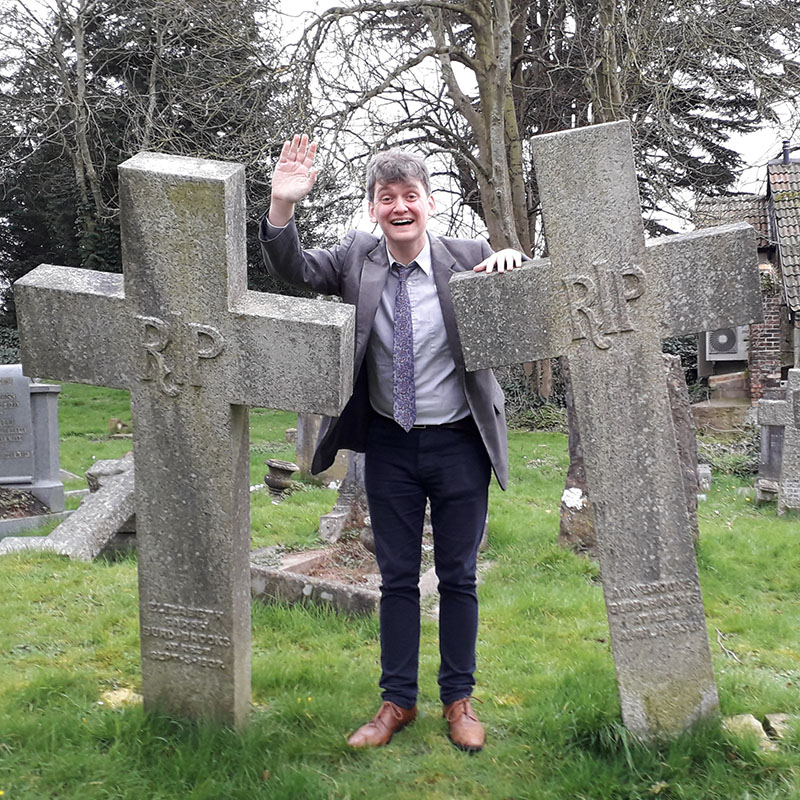 Photo of Peter Fleming smiling and waving stood between two large stone crosses in a graveyard