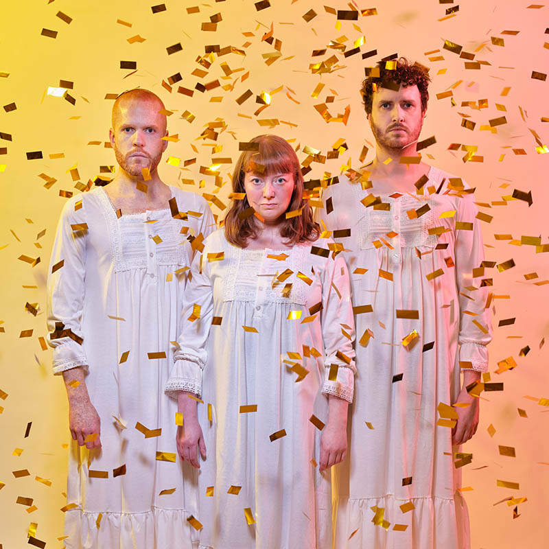 Photo of the three members for Tarot in white nightgowns with gold pieces of foil raining down on them