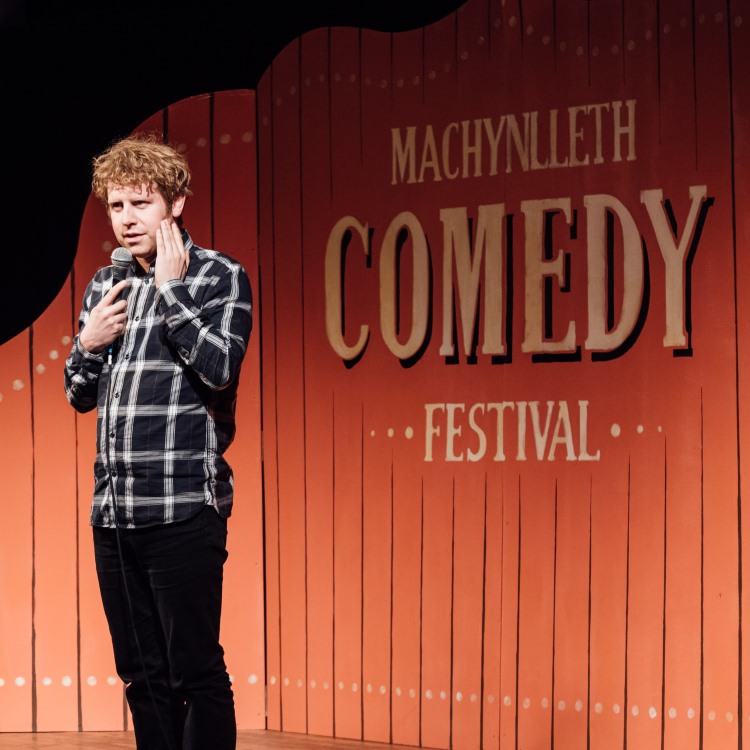 Photo of Josh Widdicombe on stage at a previous Mach Comedy Festival show in front of a festival backdrop