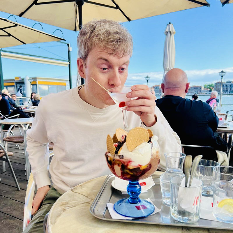 Picture of James Acaster sat under a parasol in an outside bar sipping a cocktail