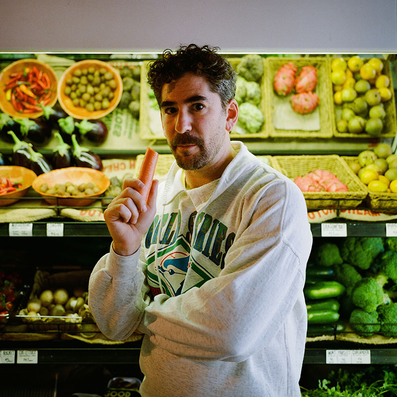 Photo of Josh Glanc stood in front of fruit & veg in a supermarket, eating a carrot