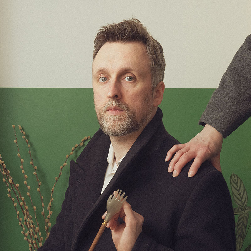 Photo of Matt Winning sat down and holding a small back scratcher in hi hand. Someone else, out of shot, is leaning on his shoulder.