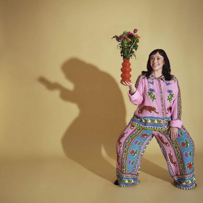 Photo of Amy Annette smiling and holding a vase in her hand. A sinister shadow is behind cast behind her looking like she is hunched over holding what looks like a phone