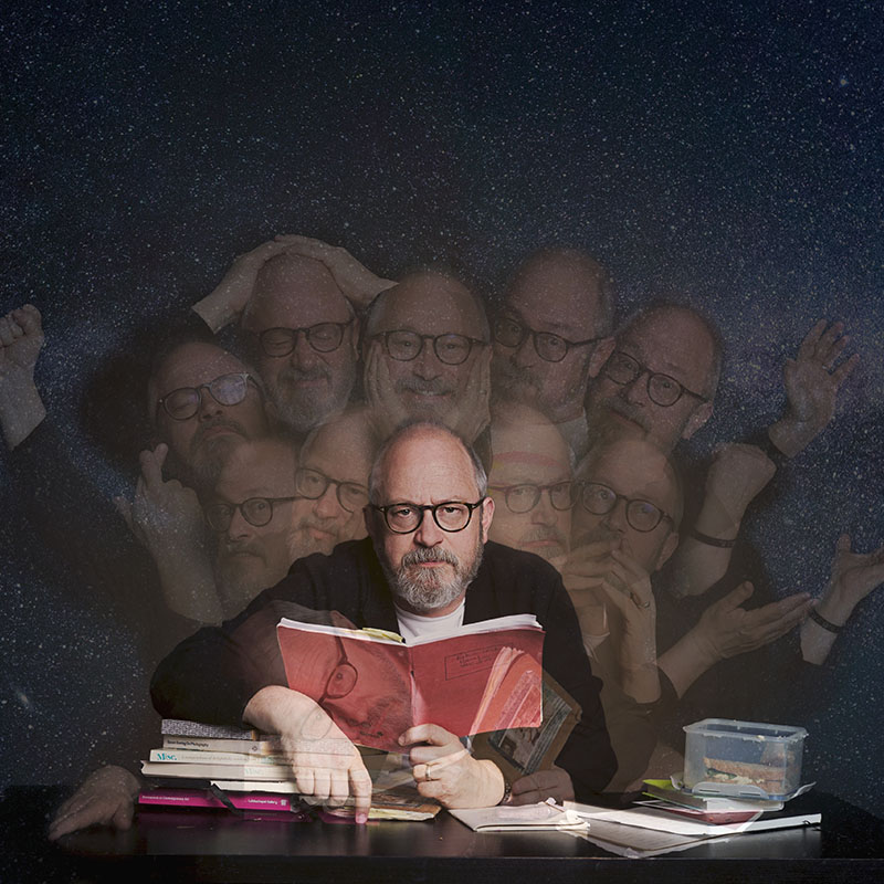 A photo of Robin Ince sat at a desk holding a school exercise book open in front of him. All around him are faint versions of him sat in the desk in various positions.