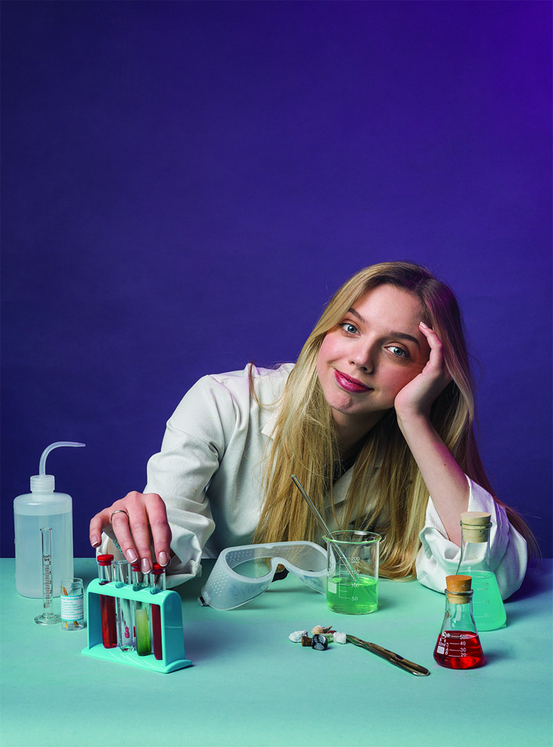 Photo of Freya McGhee sat at a table in a lab coat with lots of science paraphernalia (test tubes etc) in front of her