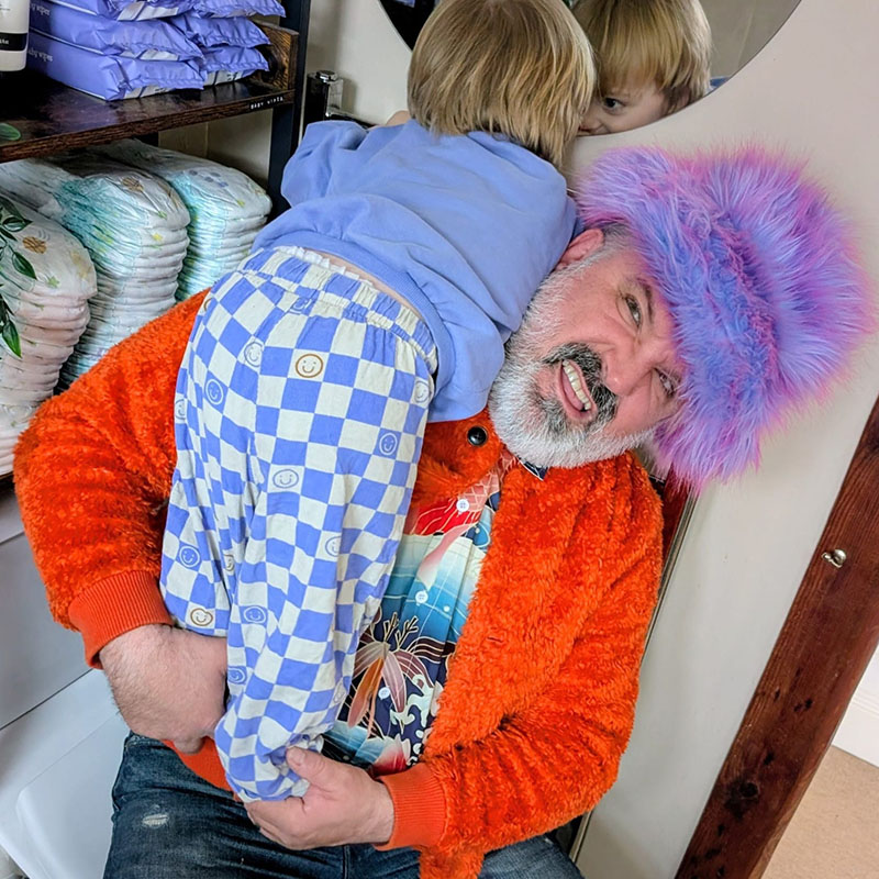 Photo of Rich Wilson in a colourful furry hat holding a toddler over his shoulder. In the backround there is a stock of nappies and wipes.