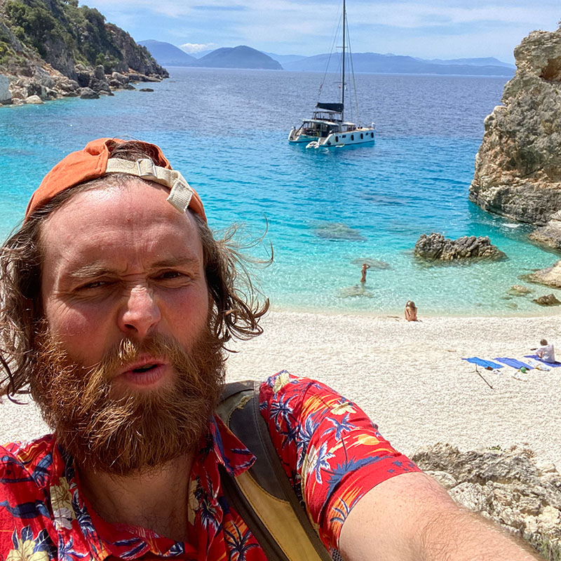Selfie of Rob Auton wearing a Hawaiian short and a baseball cap backwards in front of a sandy beach, clear blue sea and a sailing boat in the distance