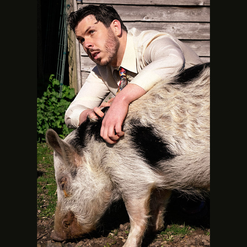 Photo of Dom McGovern kneeling down with his arms over a pig eating some grass