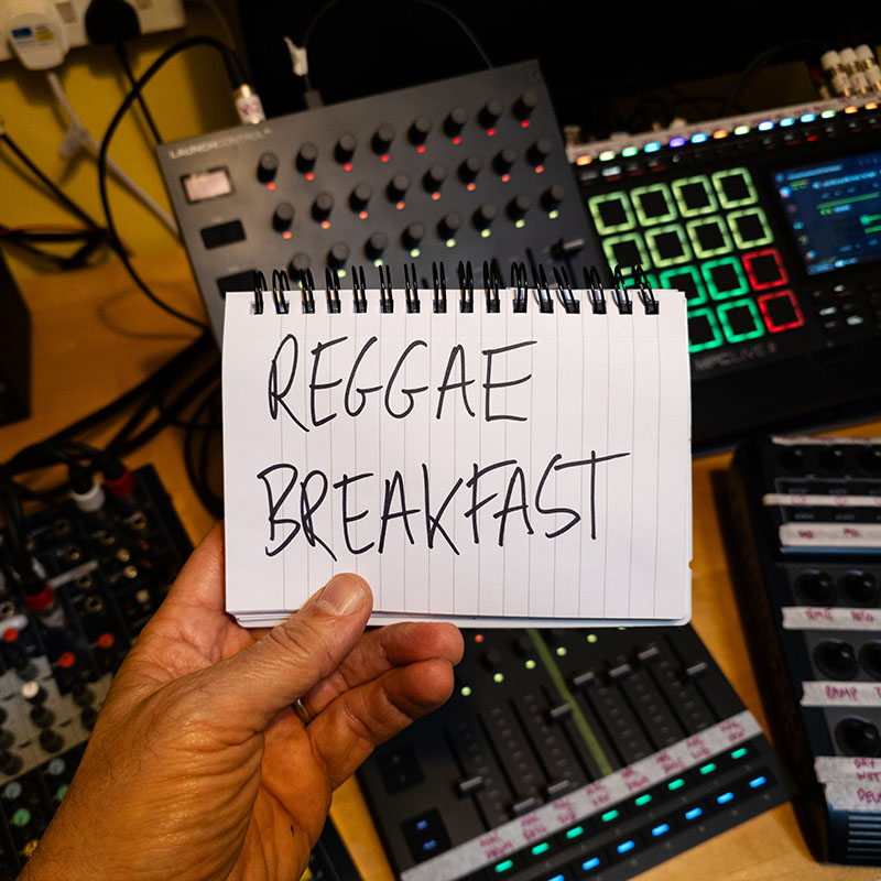 A photo of REGGAE BREAKFAST hand written onto a notepad and held over a desk full of mixers, samplers and other music production equipment