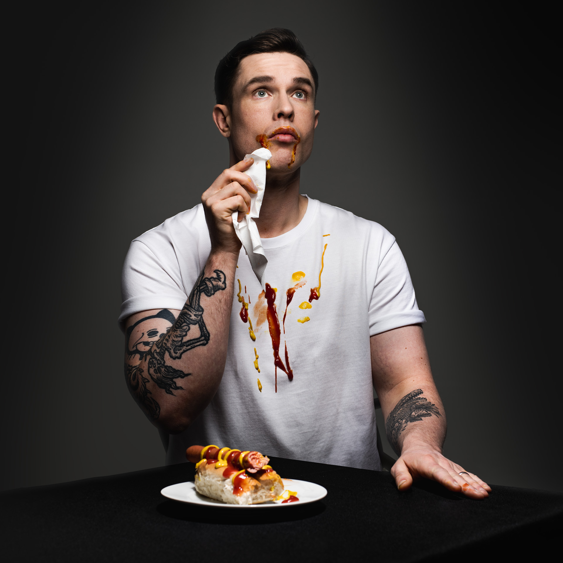 Picture of Ed Gamble with a hot dog on a plate in front of him, dabbing the corner of his mouth with a napkin with sauce spilt all down the front of his white t shirt