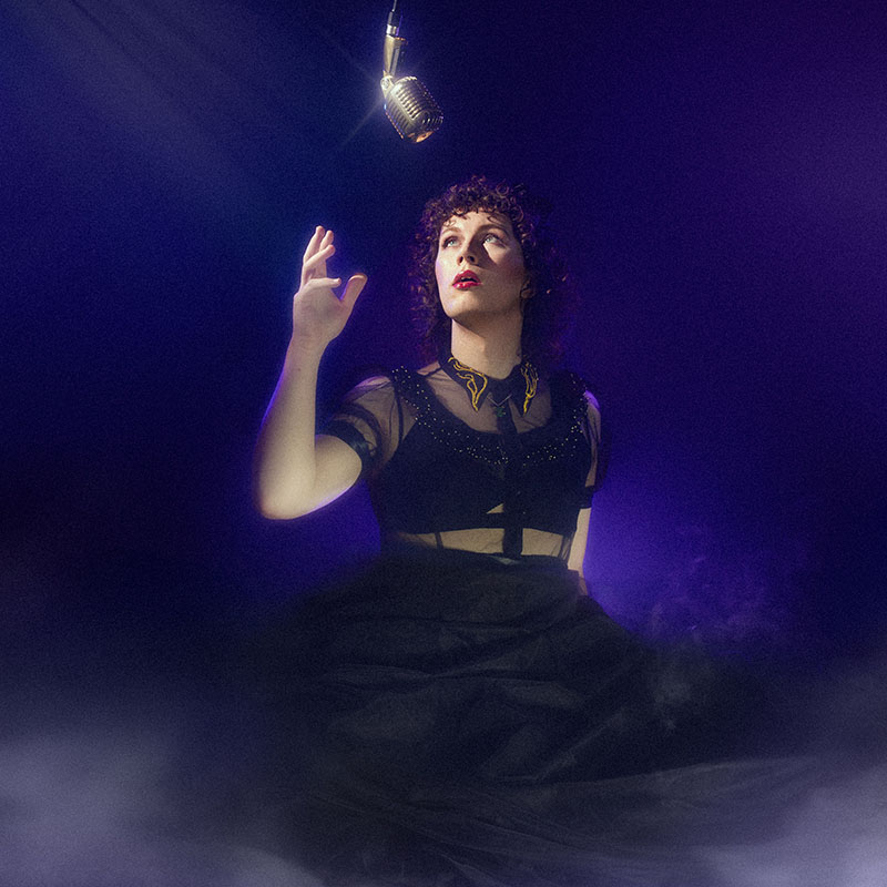 Photo of Sam Nicoresti in a black dress reaching up to an old style microphone coming down from the sky with a thin layer of smoke on the ground