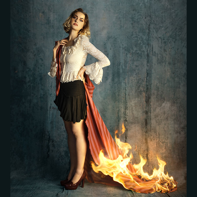 Photo of Amy Matthews wearing a black skirt and white top. She has a long piece of material wrapped around her which is trailing onto the floor. The bottom of the material is on fire very close to her legs.