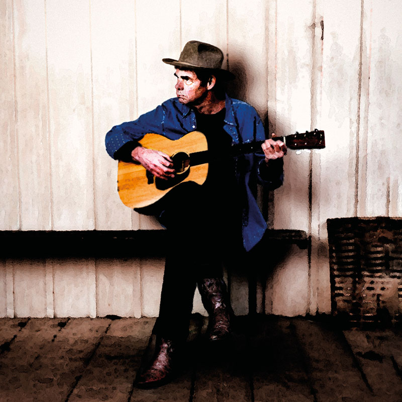 Photo of Rich Hall sat on a bench, playing his guitar