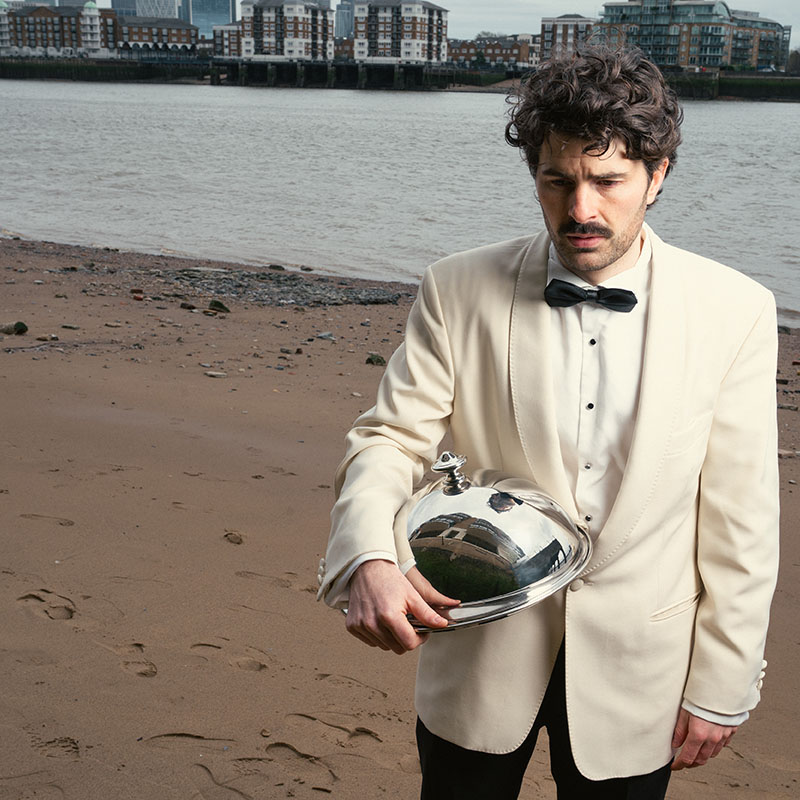 Photo of Paddy Young on a beach, wearing a tuxedo and holding a cloche under his arm