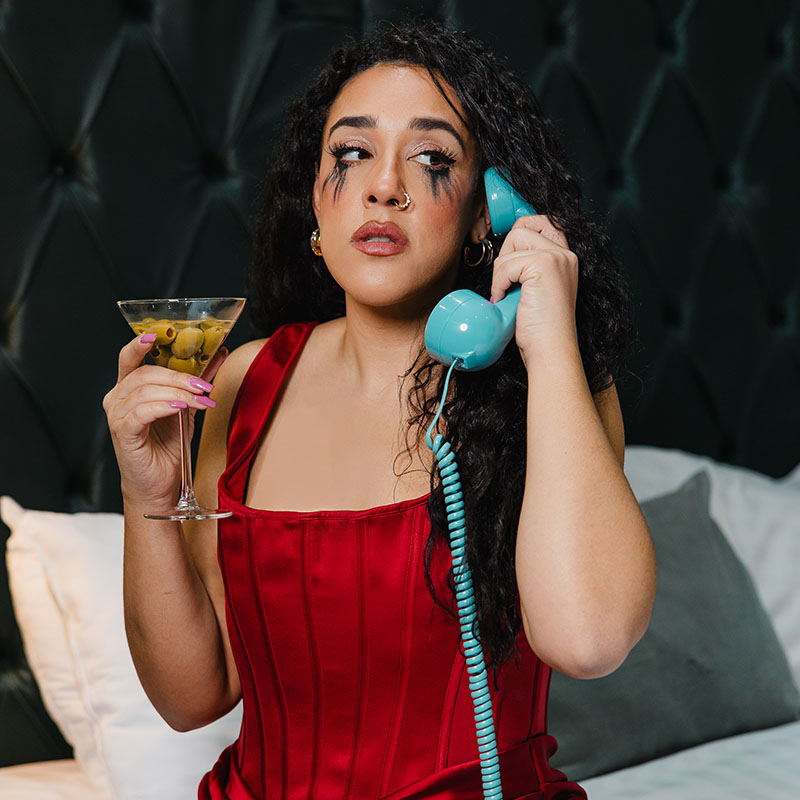 Photo of Mel Owen sat down with an old phone handset to her ear. She is wearing a dress and has mascara running down her face. In her other hand, she has a martini glass full of olives.